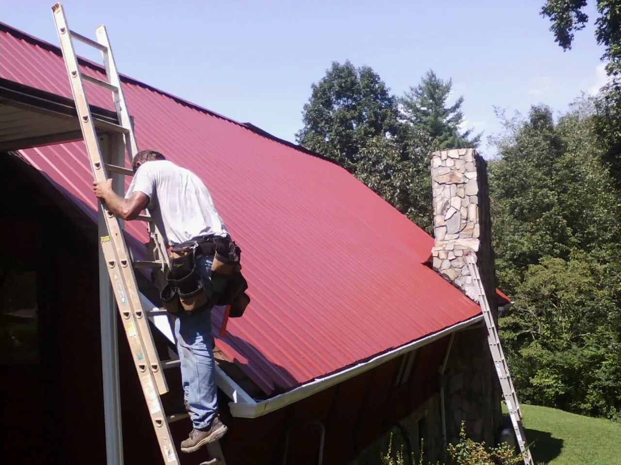 Roofer on a ladder beside a red metal roof and stone chimney — Soffit & Fascia Repair by Roofing Brothers in Bolinas